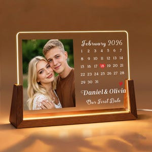 May include: A personalized illuminated acrylic photo frame with a wooden base. The frame displays a couple's photo and a calendar for February 2026, highlighting the 18th. Text reads "Daniel & Olivia, Our First Date."