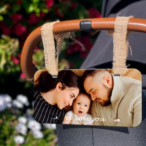 May include: A wooden photo frame with a picture of a family of three. The frame is hanging from a stroller handle with two brown fabric straps. The photo shows a woman, a man, and a baby. The text "We love you" is written on the frame.