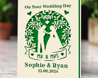 May include: A white card with green cutouts of a tree and a couple holding hands. The text on the card reads "On Your Wedding Day", "Mr & Mrs", "Sophie & Ryan", and "12.08.2024".