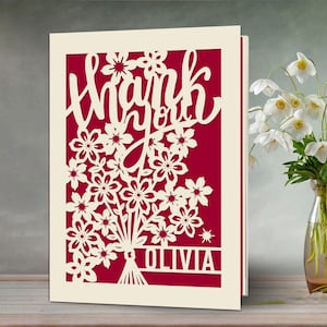 May include: A white card with a red background and a cut-out design of flowers and the words "Thank you" with the name "Olivia" at the bottom.