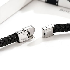 Op de afbeelding: Een zwarte gevlochten leren armband met een zilveren sluiting. De sluiting is gegraveerd met de woorden "Stainless Steel".
