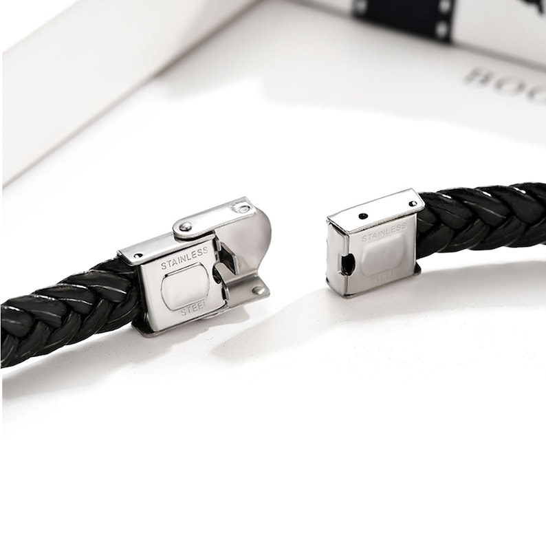 Op de afbeelding: Een zwarte gevlochten leren armband met een zilveren sluiting. De sluiting is gegraveerd met de woorden "Stainless Steel".