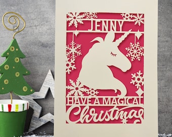 May include: A white card with a pink background and a cut-out design of a unicorn with snowflakes. The card says "Jenny Have a Magical Christmas".