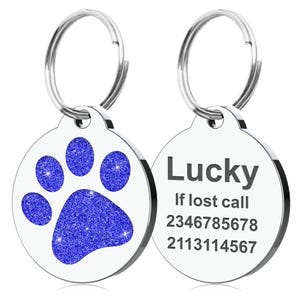 Op de afbeelding: Twee ronde, zilveren huisdier-ID-tags. E&eacute;n tag heeft een blauwe glitter pootafdruk. De andere tag heeft het woord "Lucky" en twee telefoonnummers. Beide tags hebben een zilveren ring voor bevestiging.