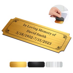 May include: Gold-colored metal plaque with a black engraved inscription that reads "In Loving Memory of David Smith 3/18/1952-7/15/2023". The plaque has a decorative border with a scalloped edge.