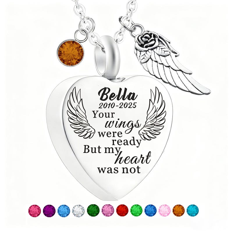 May include: A silver heart-shaped pendant necklace with the inscription "Bella 2010-2025 Your wings were ready But my heart was not." The necklace includes a charm with a rose and wing design, and a small orange gemstone. A row of colorful gemstones is at the bottom.
