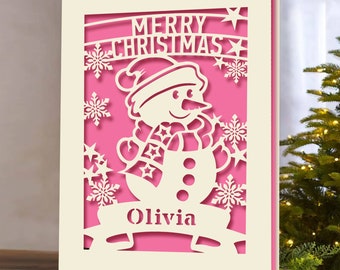 May include: A white card with a pink background and a cut-out design of a snowman with a scarf and a hat. The card says "Merry Christmas" and "Olivia" on it.