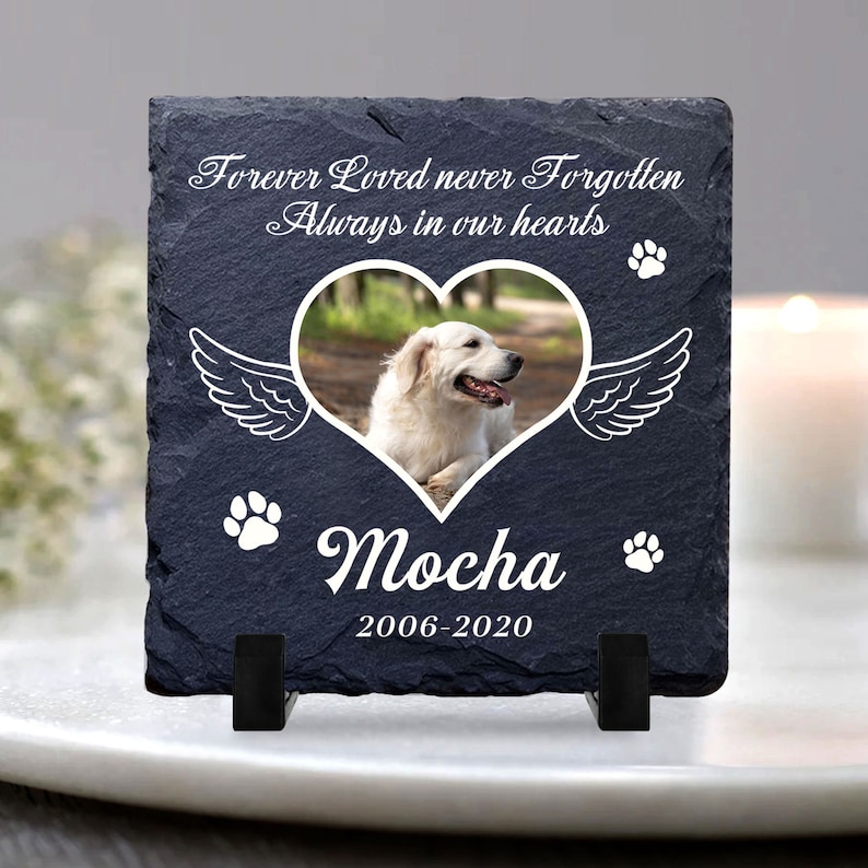 Puede incluir: Una placa conmemorativa de pizarra negra con un marco de foto en forma de coraz&oacute;n blanco, una huella de pata blanca y el texto "Forever Loved never Forgotten Always in our hearts". El marco de la foto contiene una imagen de un perro blanco. La placa tambi&eacute;n tiene el texto "Mocha 2006-2020" y dos huellas de patas blancas.