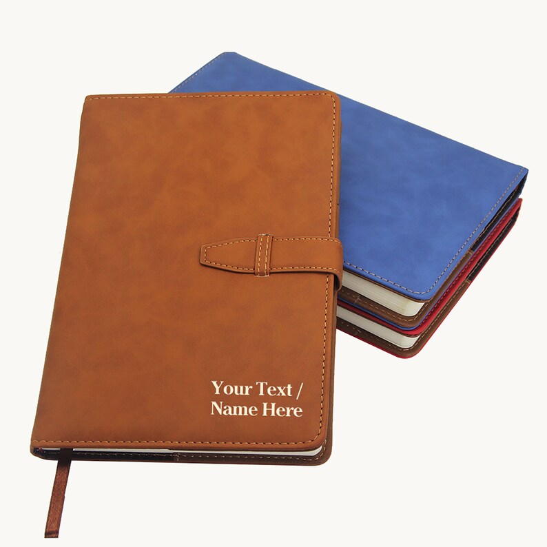 May include: A brown leather-bound journal with a strap closure and the text "Your Text / Name Here" embossed on the front cover. The journal is shown next to a stack of journals in various colors.