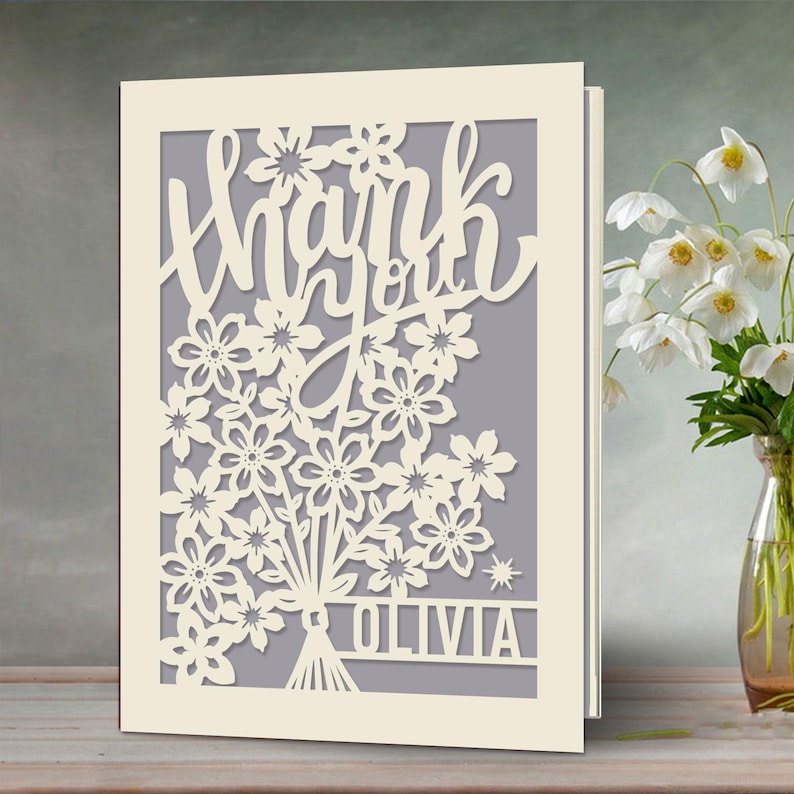 May include: A white card with a cut-out design of flowers and the words "Thank you" in a cursive font. The name "Olivia" is at the bottom of the card.