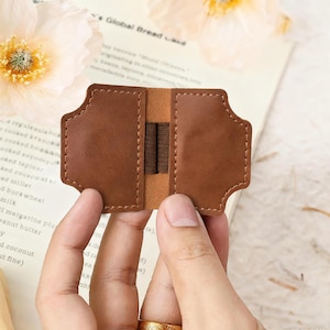 May include: A brown leather bookmark with a unique shape, featuring a central elastic band. The bookmark is held open, showcasing its interior design. The overall aesthetic is simple and elegant, suitable for book lovers.