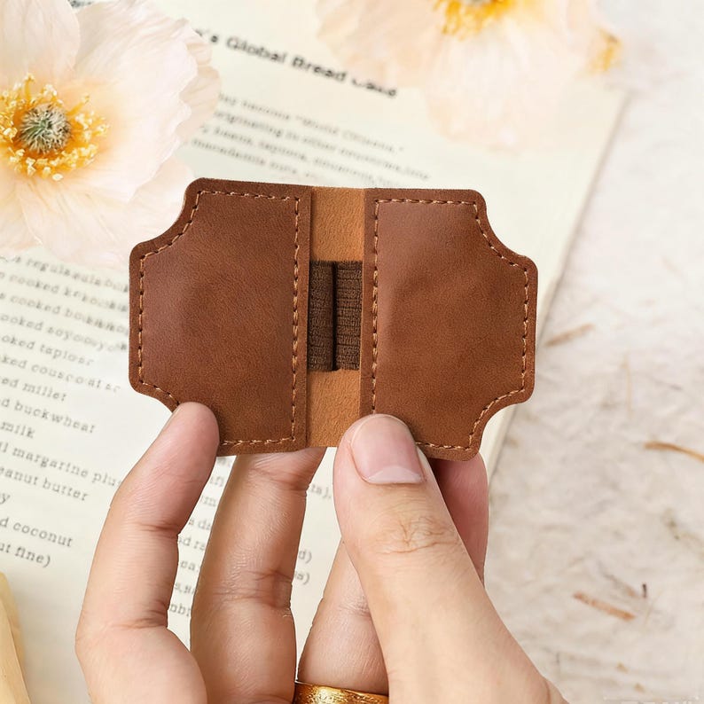 May include: A brown leather bookmark with a unique shape, featuring a central elastic band. The bookmark is held open, showcasing its interior design. The overall aesthetic is simple and elegant, suitable for book lovers.