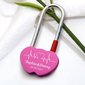 May include: Pink heart-shaped padlock with a silver shackle and a red accent. The padlock has a white heartbeat design with a heart in the middle and the text "Sophia & Danny 05.08.2024".