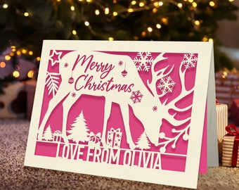 May include: A white paper Christmas card with a pink background. The card features a cut-out design of a reindeer with snowflakes and a Christmas tree. The text "Merry Christmas" and "Love from Olivia" is printed on the card.