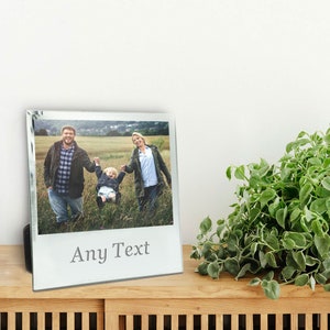 May include: A clear glass photo frame with a family photo inside. The photo shows a family of three holding hands and swinging a child in a field. The text "Any Text" is etched at the bottom of the frame.