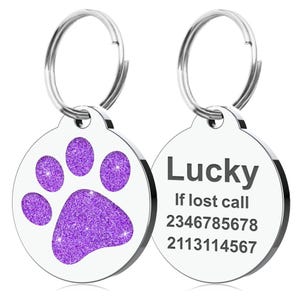 Op de afbeelding: Twee ronde, zilveren hondenpenningen. E&eacute;n penning heeft een paarse glitter pootafdruk. De andere penning heeft de naam "Lucky" en twee telefoonnummers. Beide penningen hebben een zilveren ring.