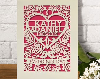 May include: A white card with a pink background and a cut-out design of a heart with the names Kathy & Daniel and the date 19/06/2019. The heart is surrounded by flowers and leaves. The text "Wedding Day" is below the heart.