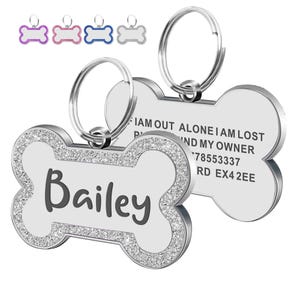 Op de afbeelding: Honden-ID-tag in de vorm van een bot met de naam "Bailey" in zwarte letters. De tag heeft een zilveren rand ingelegd met kleine, sprankelende kristallen. De tag bevat een zilveren ring voor bevestiging en de tekst "I AM OUT ALONE I AM LOST FIND MY OWNER 78553337 RD EX42EE".