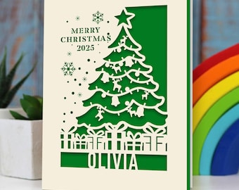 May include: A cream-colored Christmas card with a green cut-out design. The design features a Christmas tree, gifts, snowflakes, and the text "MERRY CHRISTMAS 2025" and "OLIVIA". The card is standing upright.