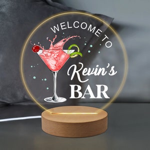May include: A wooden base with a round, illuminated sign that says "Welcome to Kevin's Bar". The sign features a cartoon illustration of a martini glass with a cherry and lime garnish.