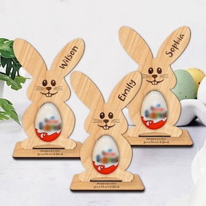 May include: Three wooden Easter bunny decorations with personalized names and a Kinder Surprise egg in the center. Each bunny has a base with the text "Happy Easter Love Mum & Dad". The names "Wilson", "Emily", and "Sophia" are engraved on the ears.
