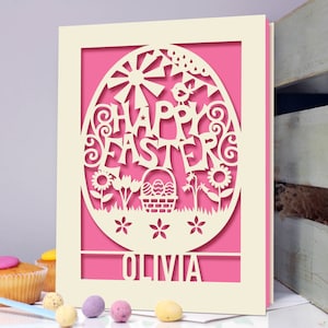 May include: A white card with a pink background and a cut-out design of an Easter egg with the words "Happy Easter" and a basket of eggs. The name "Olivia" is written at the bottom of the card.