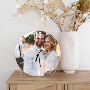 May include: A round, clear glass clock with a photo of a couple in front of a blurred background. The clock face has black hands and white numbers. The text "The Smiths EST. 2019" is printed on the clock face.