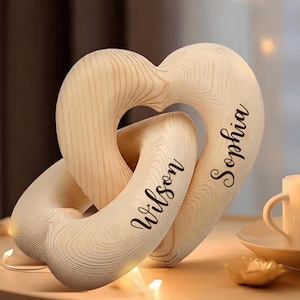 May include: A decorative wooden sculpture consisting of two interlocking heart shapes with a light natural wood grain finish. The names Wilson and Sophia are engraved in a dark cursive script on the front surfaces of the two heart forms.