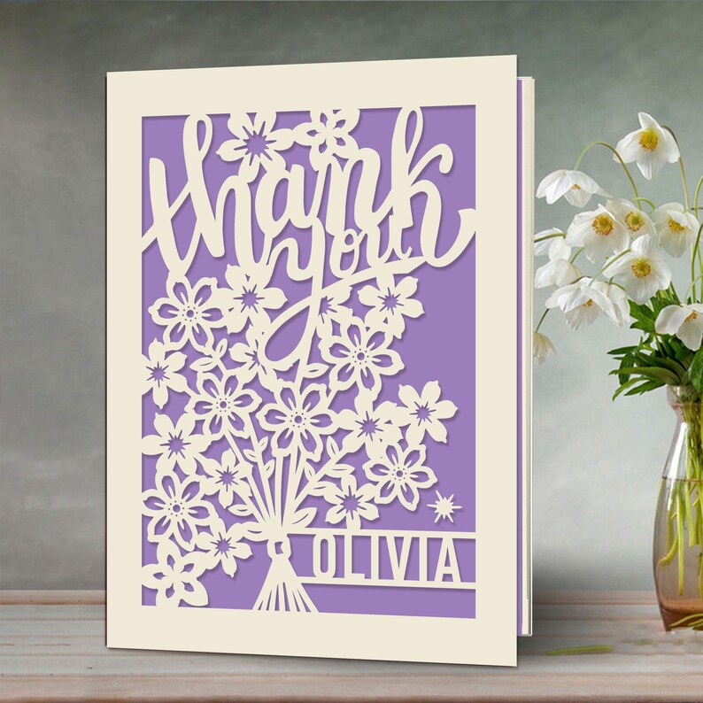 May include: A white card with a cut-out design of flowers and the words "Thank you" in a cursive font. The card has a purple background and the name "Olivia" is written at the bottom.