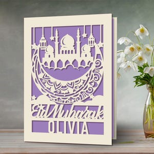 May include: A white card with a cut-out design of a mosque and crescent moon on a purple background. The text "Eid Mubarak Olivia" is at the bottom of the card.