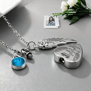 May include: A silver-colored memorial necklace with a heart-shaped pendant and a blue gemstone charm. The heart pendant has engraved text and wing designs. A small photo and flowers are in the background.