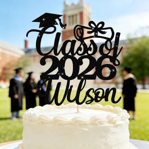 May include: A black glitter cake topper with the words "Class of 2026 Wilson" and a graduation cap and diploma design. The topper is placed on a white frosted cake, with graduates in the background.