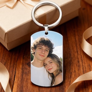 May include: A silver metal keychain with a rectangular pendant featuring a color photograph of a couple. The keychain has a circular ring at the top. The background includes a gift box and ribbon.