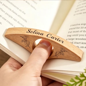 May include: A light-colored wooden thumb page holder featuring a central circular opening for a thumb. The surface is engraved with the name Selina Carter in a cursive script, flanked by two delicate line-art illustrations of morning glory flowers.