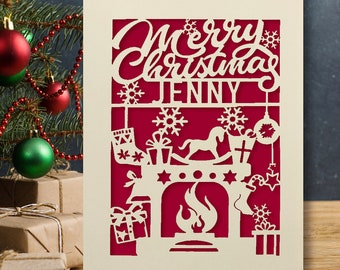 May include: A white card with a red background and a cut-out design of a fireplace with a rocking horse, stockings, and presents. The text "Merry Christmas Jenny" is at the top of the card.