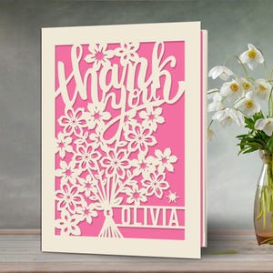 May include: A white card with a pink background and a cut-out design of flowers and the words "Thank you" with the name "Olivia" at the bottom.