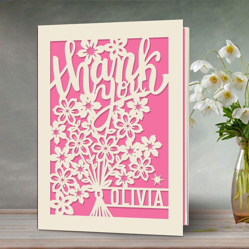 May include: A white card with a pink background and a cut-out design of flowers and the words "Thank you" with the name "Olivia" at the bottom.