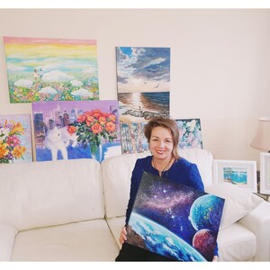 May include: A woman in a blue shirt is holding a painting of a blue planet with stars and a red planet in the background. The painting is surrounded by other paintings on the wall behind her.