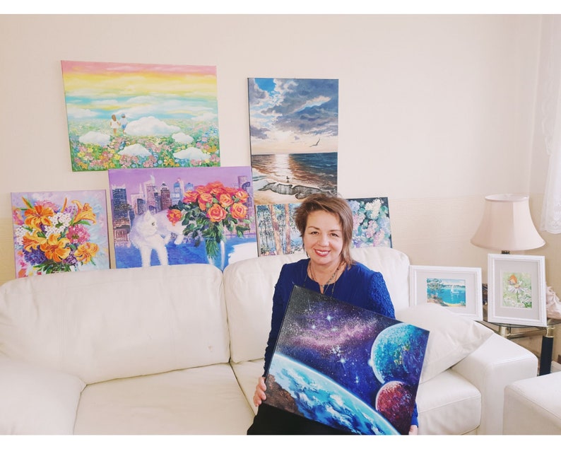 May include: A woman in a blue shirt is holding a painting of a blue planet with stars and a red planet in the background. The painting is surrounded by other paintings on the wall behind her.