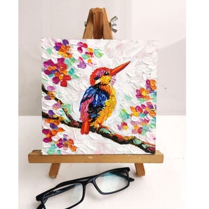 Puede incluir: Pintura al óleo texturizada de un pájaro colorido posado en una rama, sobre un fondo blanco. El pájaro está pintado con colores brillantes, incluyendo azul, naranja y rojo. La obra se exhibe en un pequeño caballete de madera.