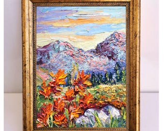 May include: Oil painting of a mountain landscape with orange wildflowers in the foreground. The artwork is framed in a gold-colored, ornate frame. The sky is a mix of blue, orange, and pink hues.