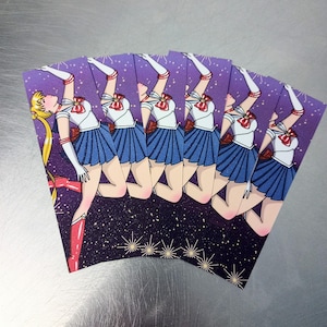 May include: A fan of six cards featuring a cartoon illustration of a character in a sailor uniform. The character is depicted with blonde hair, a white top, blue skirt, and red accents, set against a purple starry background.
