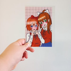 Puede incluir: Una colorida impresión de estilo anime con dos chicas. Una chica tiene el pelo corto y naranja y hace un corazón con las manos. La otra tiene el pelo largo y naranja y un lazo blanco. Llevan tops rojos y faldas azules, sobre un fondo de cuadrícula rosa.
