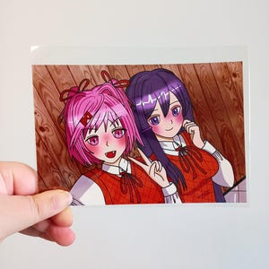 Puede incluir: Una colorida impresión de estilo anime con dos personajes femeninos. Una tiene el pelo rosa y la otra morado. Llevan chalecos rojos sobre camisas blancas. El fondo es un patrón de vetas de madera.