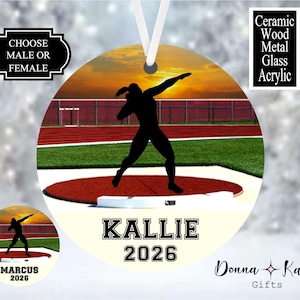 May include: A round ornament featuring a silhouette of a shot put athlete on a track at sunset. The text reads CHOOSE MALE OR FEMALE, CERAMIC WOOD METAL GLASS ACRYLIC, KALLIE, 2026, MARCUS, 2026, and Donna Kay Gifts. It includes a white ribbon for hanging.