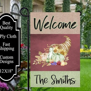 May include: A welcome garden flag with a fall theme. The flag is divided into three sections. The top section is a beige burlap with the word "Welcome" in black script. The middle section is a burgundy background with a cornucopia overflowing with gourds, apples, grapes, and a sunflower. The bottom section is a green burlap with the text "The Smiths" in black script.