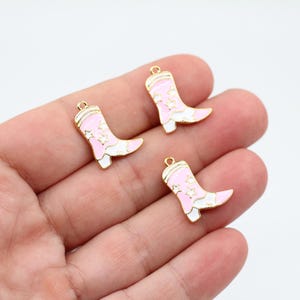 May include: Three gold-colored cowboy boot charms with pink enamel and white accents. The boots have a star design and are shown against a white background. Each charm has a small loop at the top.