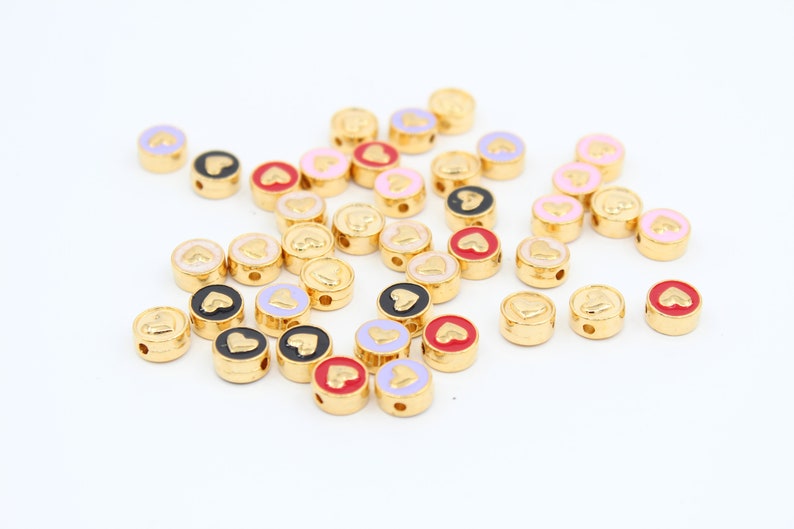 May include: A collection of gold-colored round beads with a heart design. The beads are in various colors, including pink, red, black, and white.