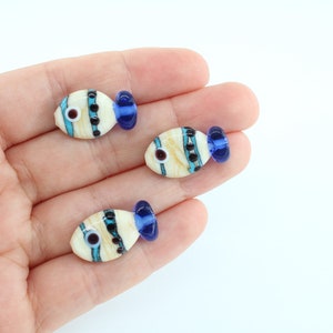 Puede incluir: Tres cuentas de pescado de vidrio con rayas blancas, azules y negras. Los peces tienen colas azules y ojos negros.