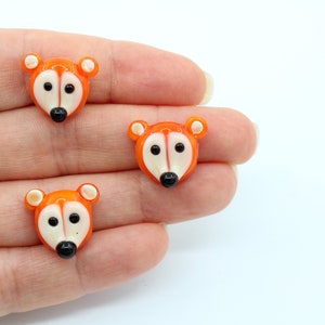 May include: Three orange glass animal buttons with white faces and black eyes. The buttons are shaped like foxes.